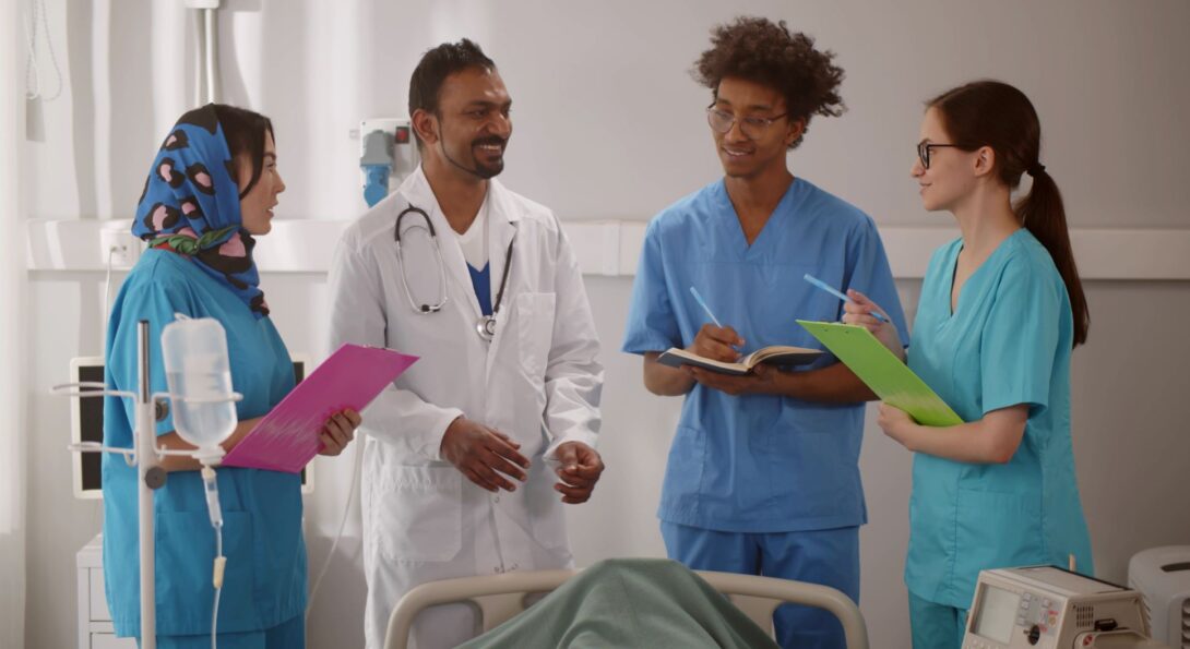 A doctor in a white coat smiles at colleagues in scrubs, who take notes.