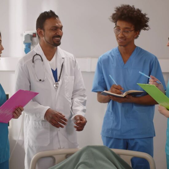 A doctor in a white coat smiles at colleagues wearing scrubs, who take notes.