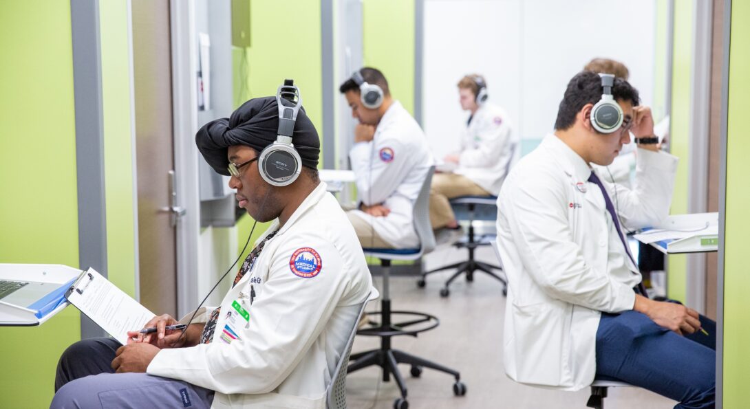 Students in white coats sit at desks with headphones on.