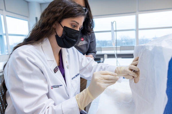 A student in a white coat practices a procedure with staff member watching.