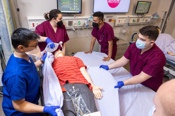 Students in scrubs and gloves transfer a mannequin to exam table