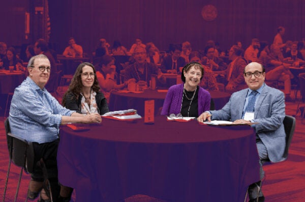 Group of four people sitting at a round table in a conference. The background is muted with red with focus just on the table.