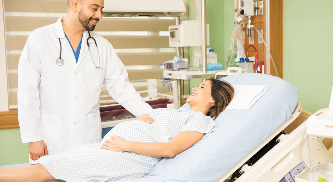 A doctor in a white coat attends to a pregnant patient in a hospital bed.